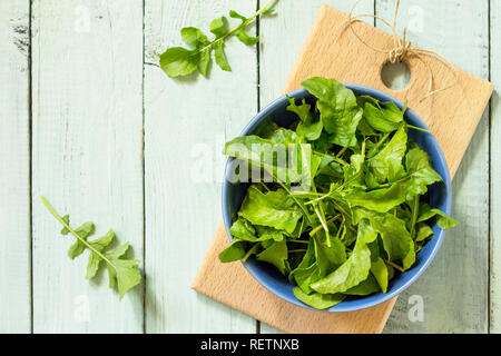 Insalata fresca foglie di rucola su rustica cucina un tavolo di legno. Vista superiore piatta sullo sfondo dei laici. Copia dello spazio. Foto Stock