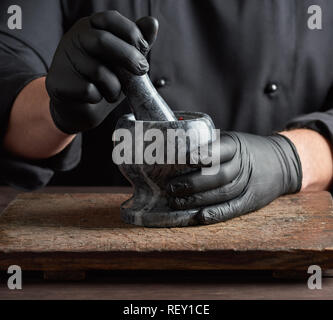 Chef in nero Guanti in lattice detiene un mortaio di pietra con una miscela di peperoni, cottura e macinazione di spezie Foto Stock
