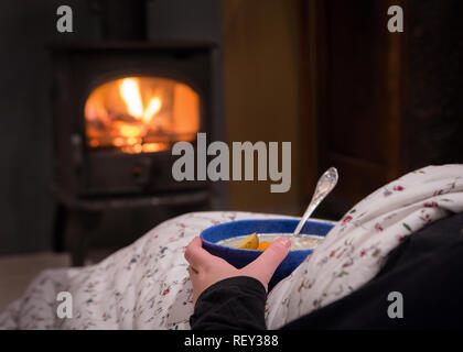 Donna con un caldo ciotola di zuppa di verdure al camino. La masterizzazione di camino con tronchi di legno che brucia in background. Luce calda, romantico, Foto Stock