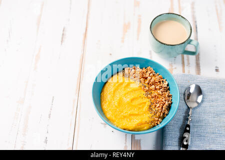 Vista superiore del frullato giallo ciotola con granola in lastra blu su bianco tavola. Spazio di copia Foto Stock