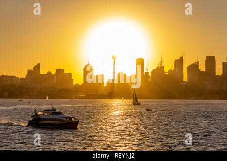 Vista del centro della città di Sydney al Tramonto con Sun impostazione disco verso il basso dietro gli edifici di una barca a vela in primo piano Foto Stock