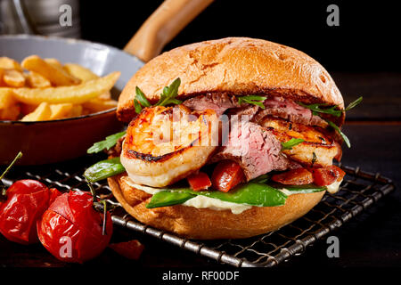 Specialty Surf and Turf burger con scampi e arrosto di manzo con rucola, petit pois e pomodoro su una griglia con un lato che serve di patatine fritte Foto Stock