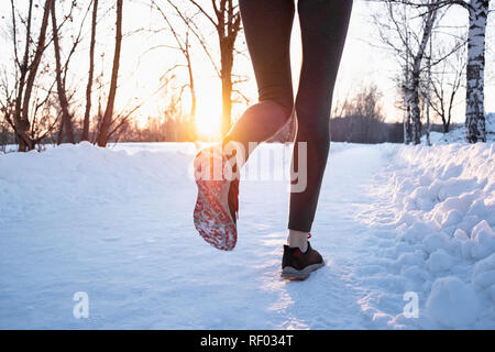 Il jogging all'aperto in inverno concetto. Le gambe di una persona di sesso femminile che corre lungo la strada innevata sulla bella giornata fredda, sparato contro il sole con lens flare Foto Stock