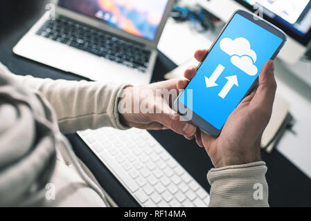 Uomo seduto alla scrivania in ufficio utilizzando il servizio cloud sullo smartphone per il trasferimento e la sincronizzazione dei dati Foto Stock