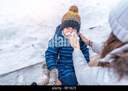 Salviette per Mom snot figlio seduto su una panchina nel parco d'inverno. Cura di una giovane madre di un bambino è un ragazzo. Il concetto della rinorrea e della congestione nasale in bambini in inverno. Foto Stock