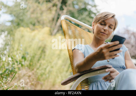 Donna seduta in giardino su una sedia con il telefono cellulare Foto Stock