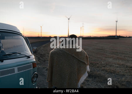 Giovane avvolto in una coperta a camper van nel paesaggio rurale con le turbine eoliche in background Foto Stock