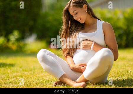 Bella donna incinta seduta sull'erba nel suo giardino Foto Stock