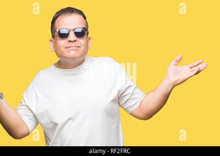 La mezza età uomo arabo wearig t-shirt bianco e occhiali da sole su sfondo isolato clueless e espressione confusa con le braccia e le mani alzate. Dubbio co Foto Stock