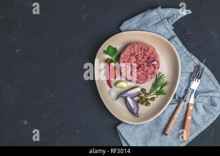 Deliziosa la tartare di manzo e gli ingredienti sulla piastra in ceramica, set di posate coltello, forchetta sulla pietra nera di calcestruzzo superficie testurizzata sfondo. Spazio di copia Foto Stock