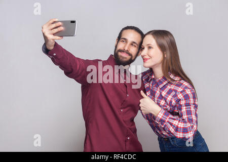 Ritratto di felice soddisfatto giovane in piedi, guardando e sorridente alla fotocamera di mobile smart phone per rendere selfie o video chiamata con il pollice in alto. interna s Foto Stock