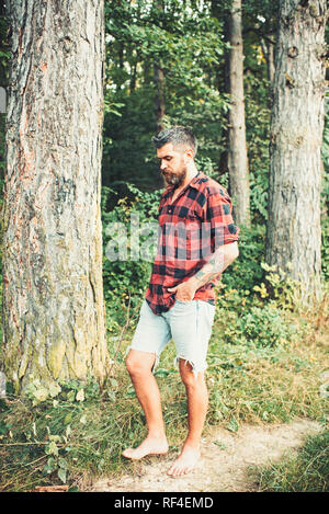 Uomo Barbuto nella foresta. Hipster con lunga barba di verde naturale paesaggio. Tourist in plaid shirt e Jean Corti il viaggio a piedi nudi. Traveler escursionismo sulla giornata di sole. La vacanza estiva concept Foto Stock