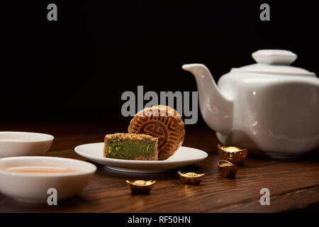 Tradizionale cinese delizioso mooncakes, teiera e oro Lingotti su un tavolo di legno isolato su nero Foto Stock