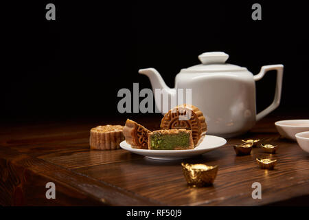 Tradizionale cinese delizioso mooncakes, teiera e oro Lingotti su un tavolo di legno isolato su nero Foto Stock