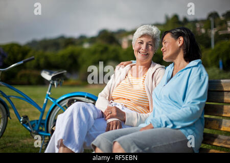 Due femmina matura amici per prendersi una pausa da ciclismo per sedersi a chiacchierare felicemente insieme su una panchina nel parco. Foto Stock