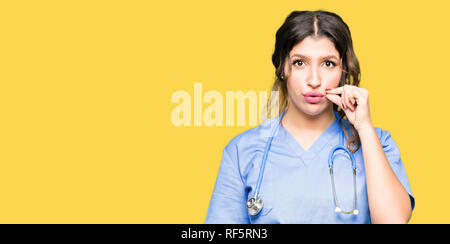 I giovani adulti medico donna che indossa uniforme medica bocca e le labbra chiuse come zip con le dita. Segreto e silenzioso, tabù parlando Foto Stock