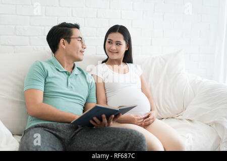 Close-up di Asian donna incinta e il marito asiatici a leggere un libro sul divano di casa. La famiglia felice in attesa il concetto del neonato. Foto Stock