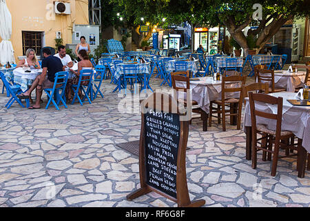 Halki Island, Grecia - 27 agosto 2018. Gruppo di persone a tradizionale taverna greca ristorante con menu inglese lavagna di Emborios square - Greco Foto Stock