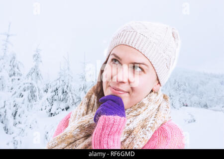 Donna che indossa il ponticello rosa e accessori invernali pensare come cercando di toccare il mento con la mano con la neve in background Foto Stock