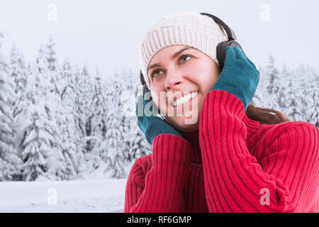 Donna sorridente godendo la musica nelle cuffie oltre il cappello bianco di toccarli con guanti verde sul paesaggio invernale sullo sfondo Foto Stock