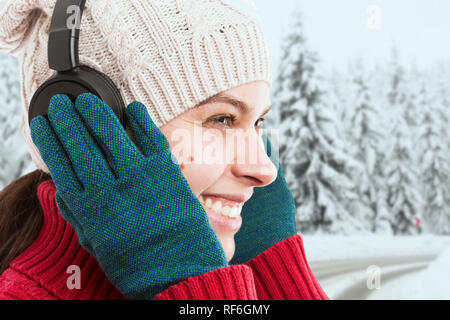 Close-up di donna toccando le cuffie con guanti verde avente felice espressione come ascoltare musica in inverno sfondo innevato Foto Stock