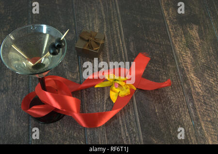 Coppetta Martini con le olive con un nastro rosso scarlatto, confezione regalo e orchidea gialla su un tavolo di legno. Close-up. Foto Stock