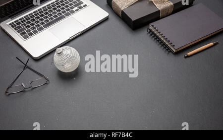 Natale a lavoro. Confezione regalo nera con nastro lucido e il computer portatile su nero scrivania da ufficio, spazio di copia Foto Stock