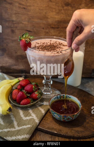 Banana, fragola, il latte e il miele su uno sfondo di legno e con un bicchiere pieno di latte shake Foto Stock