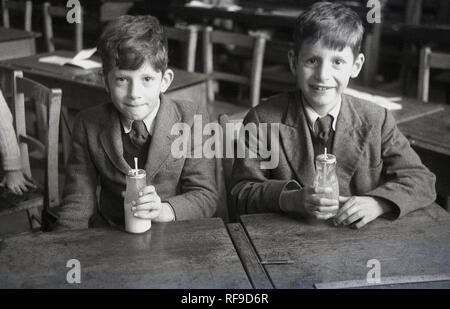 1956, storico, due scolari, eventualmente gemelli, alle loro scrivanie con bottiglie di latte alle scuole, Inghilterra, Regno Unito. Nel post-conflitto in Gran Bretagna, il latte è stato un comune a metà mattina merenda per bambini della scuola primaria dando loro nutrienti essenziali e li aiuta a mantenere idratata tra bearkfast e pranzo. Molti ci è piaciuto in inverno quando il latte era freddo e refrigerate, ma in primavera e in estate quando era spesso caldo a causa della mancanza di frigoriferi, non era così bella. Foto Stock