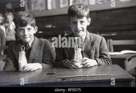 1956, storico, due scolari, eventualmente gemelli, alle loro scrivanie con bottiglie di latte alle scuole, Inghilterra, Regno Unito. Nel post-conflitto in Gran Bretagna, il latte è stato un comune a metà mattina merenda per bambini della scuola primaria dando loro nutrienti essenziali e li aiuta a mantenere idratata tra bearkfast e pranzo. Molti ci è piaciuto in inverno quando il latte era freddo e refrigerate, ma in primavera e in estate quando era spesso caldo a causa della mancanza di frigoriferi, non era così bella. Foto Stock