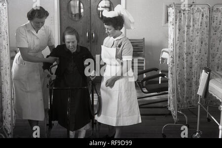 Degli anni Cinquanta, storico, due infermieri femminile aiutando un ferito lady paziente in una sedia di mobilità muoversi all'interno di un ospedale, England, Regno Unito Foto Stock