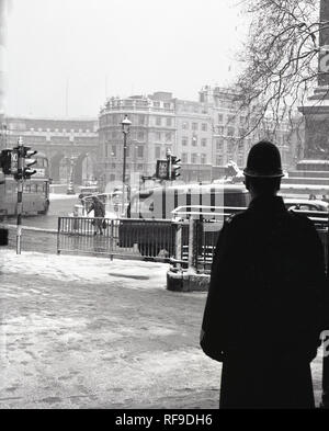 Anni sessanta, un poliziotto britannico in piedi su un marciapiede guardando un nevoso, invernale Trafalgar Square, London, England, Regno Unito Foto Stock