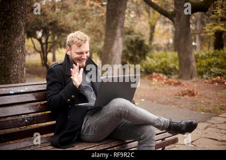 Un giovane laughing man, seduti su una panchina nel parco pubblico, utilizzando laptop, parlare su Internet, video chat o chiamata, potrebbe essere con la famiglia, amico. gestu Foto Stock