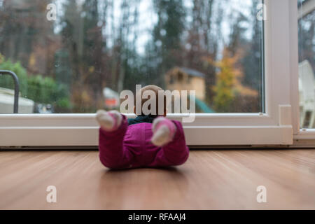 Baby girl giacente su legno soggiorno piano guardando fuori dalla finestra della porta. Foto Stock