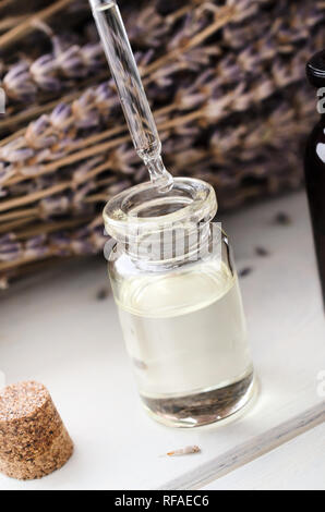 Chiusura del flaconcino di vetro trasparente con pipetta sospesa al di sopra, gocciolamento dell'olio. Gambi di erba Lavanda fiori in background. Flacone color ambra e sughero su entrambi i Foto Stock