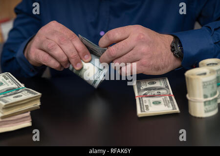 Imprenditore conteggiare denaro in rotoli e le pile in una vista ravvicinata delle sue mani di rotolamento delle banconote in USD su una tavola nera Foto Stock