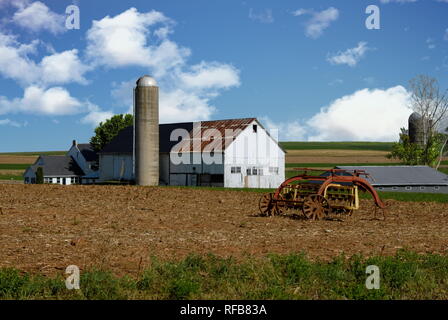 Fattoria Amish con il vecchio arrugginito Vintage Farm Equipment impostazione in campo con fienile e agriturismo in una giornata di sole Foto Stock