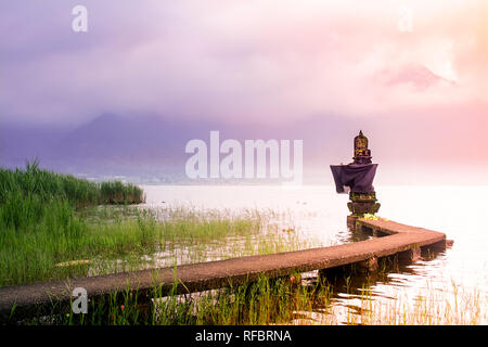 Piccolo tempio indù sul lato lago con ponte di collegamento durante il Sunrise a Bali il nuvoloso e nebbioso giorno con la montagna come sfondo. Foto Stock