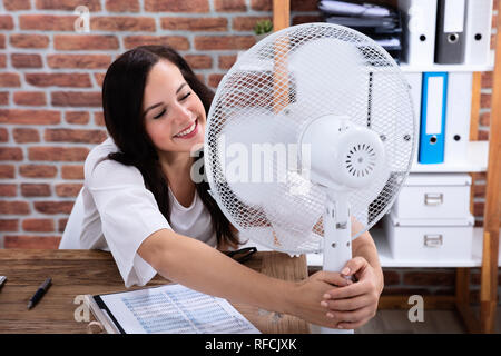 Sorridente giovane donna godendo brezza con ventola elettrica in ufficio Foto Stock