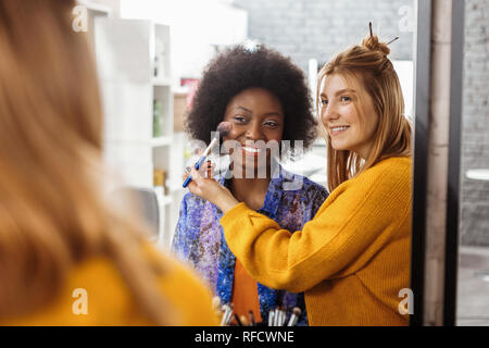 Biondo stilista sorridente cercando su un nuovo blusher swatch sulla di carnagione scura modello Foto Stock