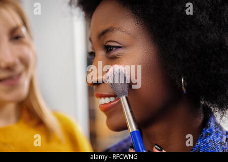 Biondo stilista sorridente cercando un nuovo look su di carnagione scura modello Foto Stock