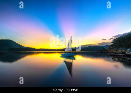 Lonely in barca a vela nel lago durante il bellissimo tramonto con raggi di sole nuvole traspare dal cielo colorato riflette il lago mistico a fine giornata Foto Stock