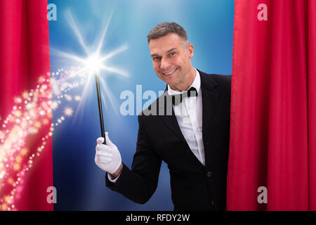 Felice maschio maturo mago tenendo la Bacchetta Magica dietro il sipario rosso Foto Stock