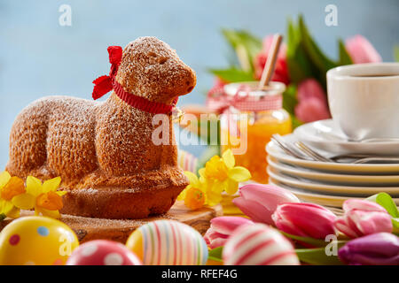 Molla di specialità torta di agnello su una tavola di Pasqua decorate con colorati uova fresche tulipani e narcisi e una pila di piastre con una tazza di caffè Foto Stock