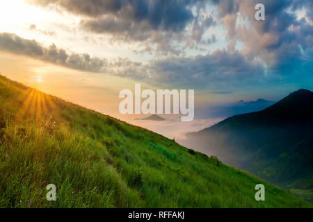 Paesaggio di montagna nelle giornate di bel tempo a sunrise. Erbaceo verde collina ripida, nebbiosa valle e monti in lontananza sotto luminose blu cielo con illuminato da salita s Foto Stock