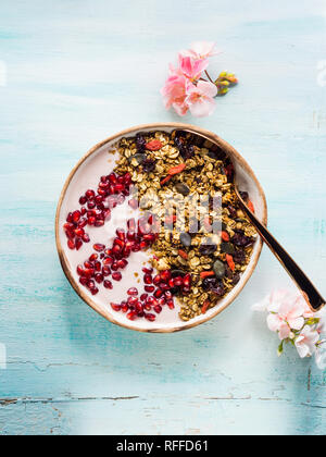 Fatti in casa la coppa di muesli con yogurt alla frutta e melograno per la prima colazione con frutta secca, semi e noci. La molla umore con fiori di colore rosa pastello su backg Foto Stock