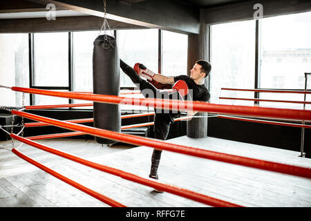 Uomo atletico calci sacco da boxe con una gamba, formazione kickboxing sul pugilato ring in palestra Foto Stock