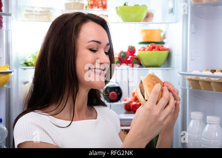 Sorridente giovane donna Guardando Burger in piedi nella parte anteriore del frigorifero Foto Stock