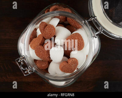 Un vaso riempito sith bianco e marrone a forma di cuore i cubetti di zucchero, al buio su un pannello di legno, laici piatta Foto Stock