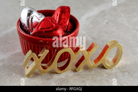 Immagine ravvicinata di rosso ramekin riempito di rosso e argento a forma di cuore ad cioccolatini. Metallo oro simbolo di baci e abbracci in primo piano. Sfondo marrone chiaro Foto Stock